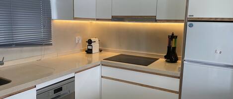 A compact kitchen with white cabinets, a built-in dishwasher, a coffee machine, and a refrigerator.