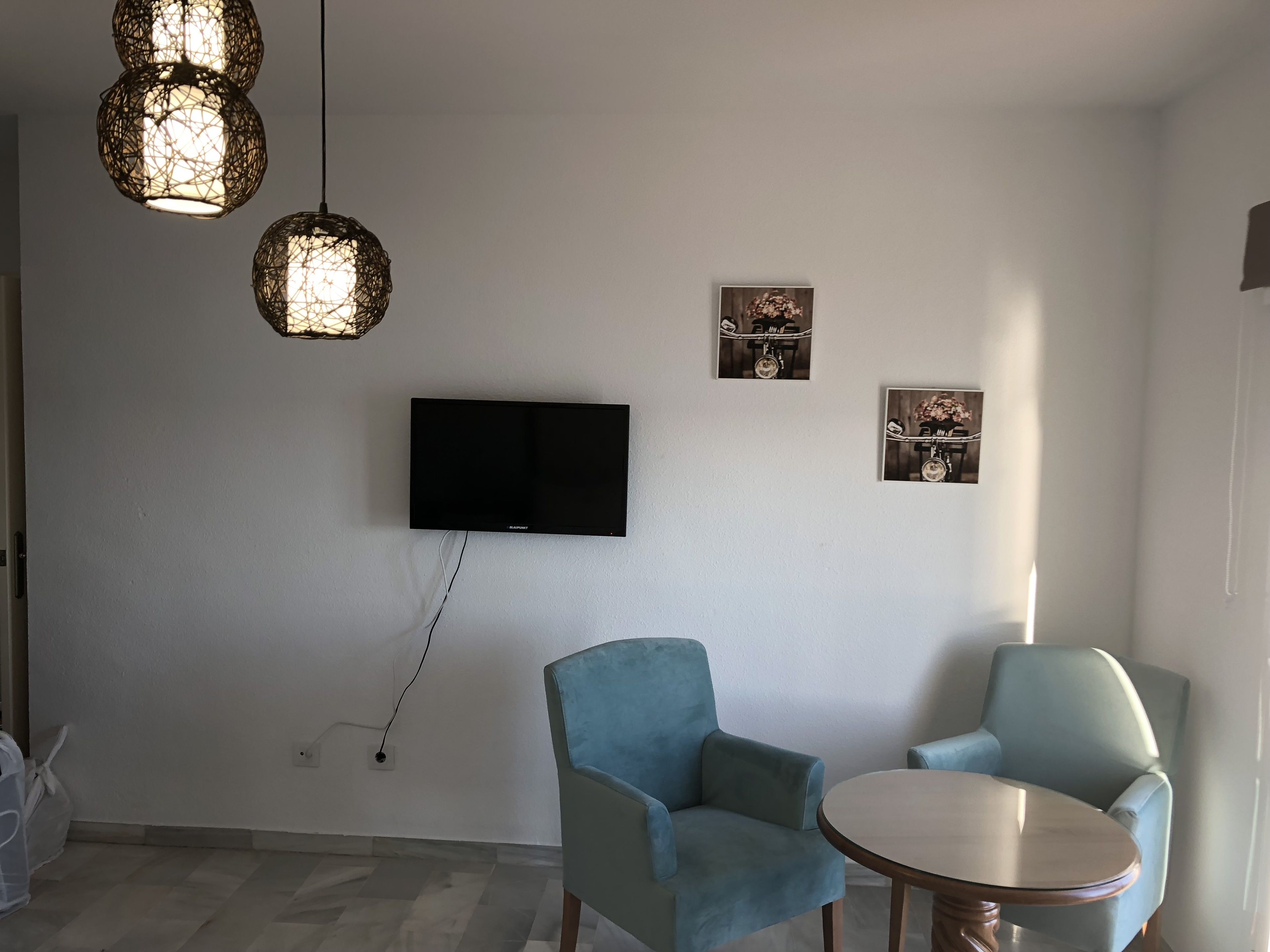 Un salon moderne doté d’un téléviseur à écran plat, de deux fauteuils bleus, d’une table ronde en bois et de trois lampes suspendues.