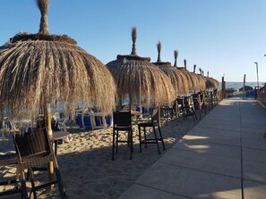 A beachside seating area with thatched umbrellas and chairs, a concrete walkway, and a clear view of the ocean. - Apartamento Balcon del Mar (Mijas)