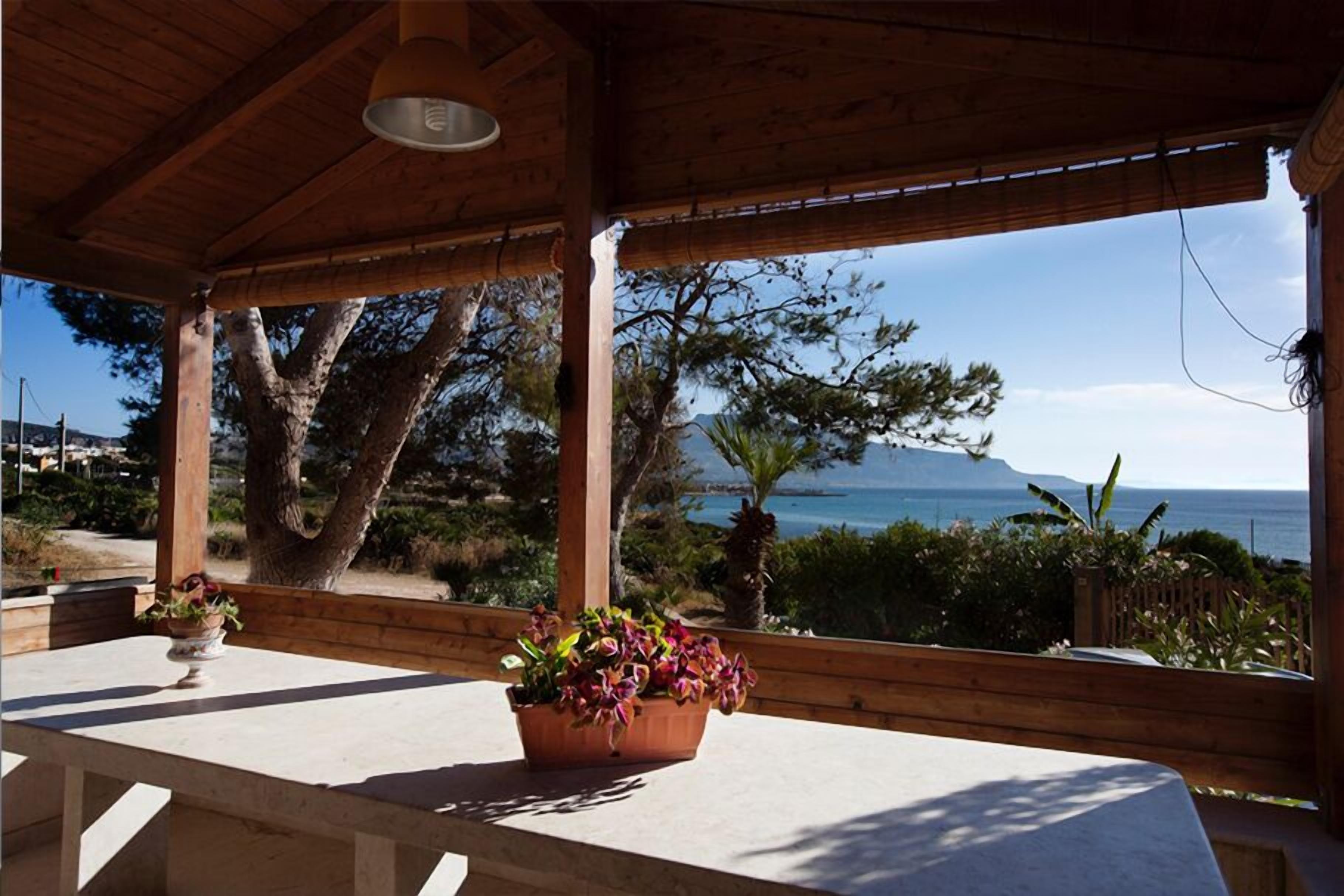 Un espace extérieur abrité, doté d’un plafond en bois, d’une table ornée d’une plante en pot, et offrant une vue sur la mer et les montagnes.