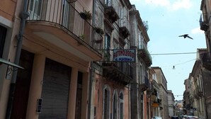 Vista de la calle con un edificio que dice "AUTONOLOGGIO G. PLUCHINO" y coches estacionados a lo largo de la vía.