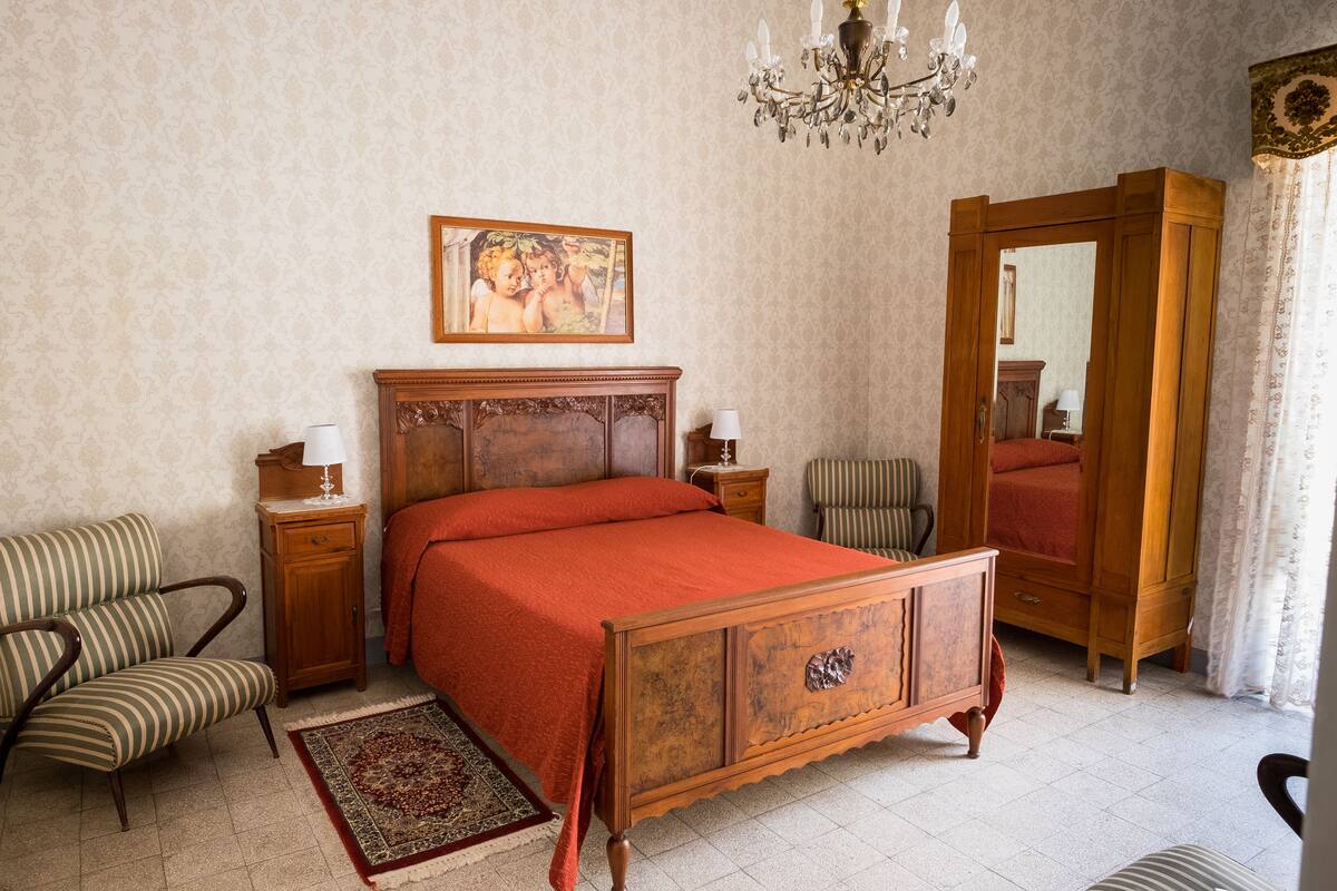 Un dormitorio con una cama de madera, una mesita de noche, una silla, un aparador, un espejo y una araña.