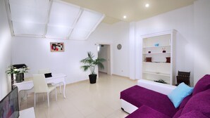 Ein modernes Wohnzimmer mit einem weißen Sofa, einem Essbereich mit Tisch und Stühlen sowie einem großen Fenster mit Jalousien.