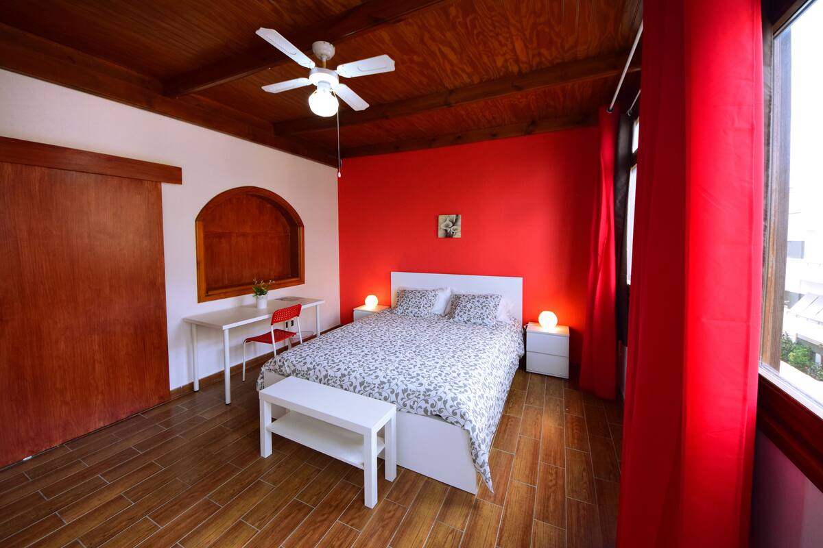 Une chambre à coucher comprenant un lit, un bureau, une chaise, un banc, un ventilateur de plafond et une fenêtre avec des rideaux rouges.
