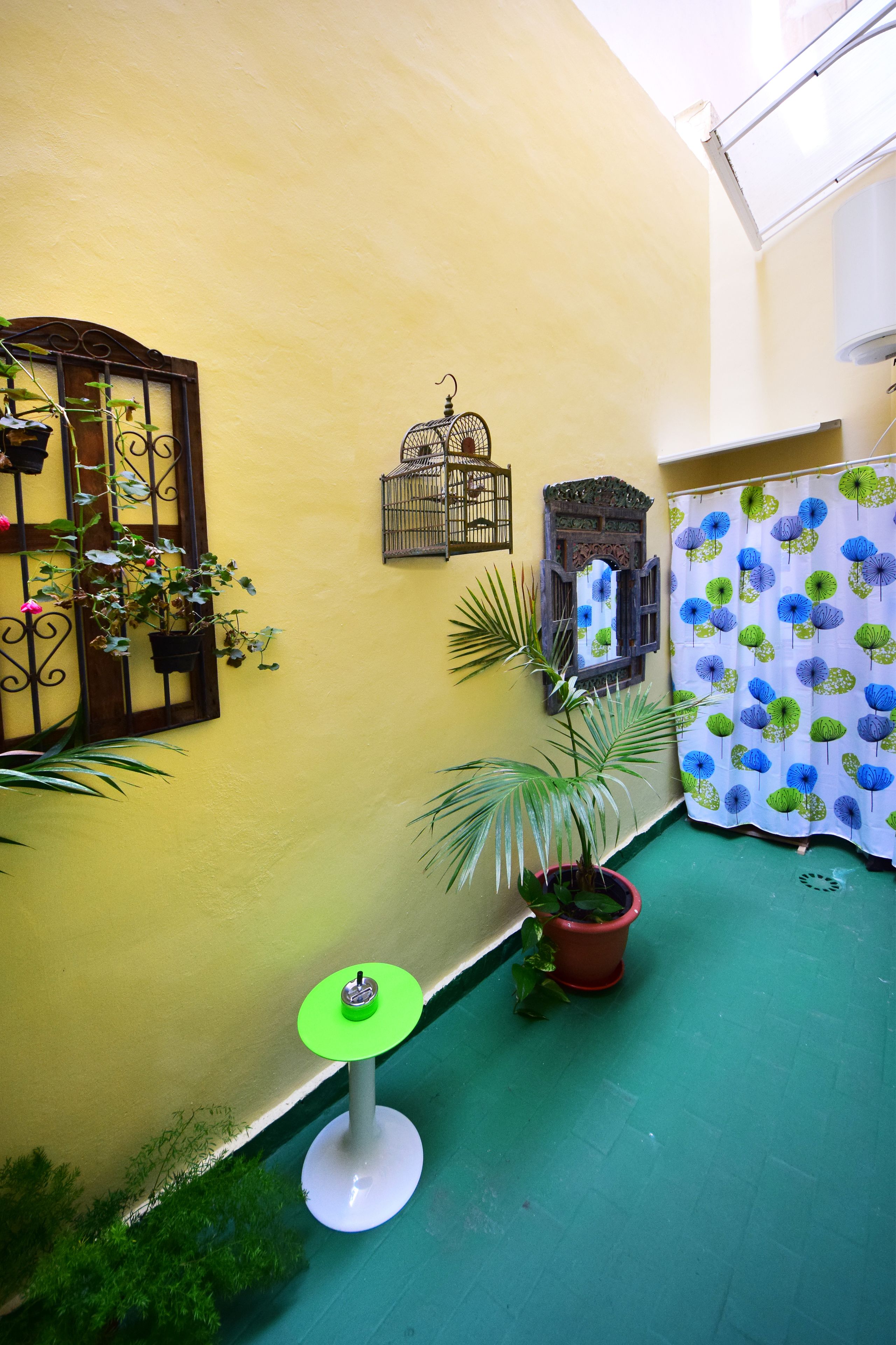 A narrow corridor with a yellow wall, a green floor, a potted plant, a small table, and a patterned curtain.