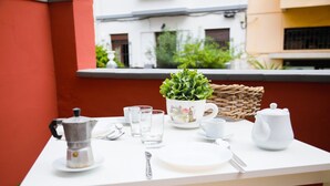 Ein weiß gedeckter Tisch mit Kaffeekanne, Gläsern, Teekanne und Tasse, mit einem Topf und einem Korbkörbchen auf einem Balkon mit roter Wand und einem Gebäude im Hintergrund.