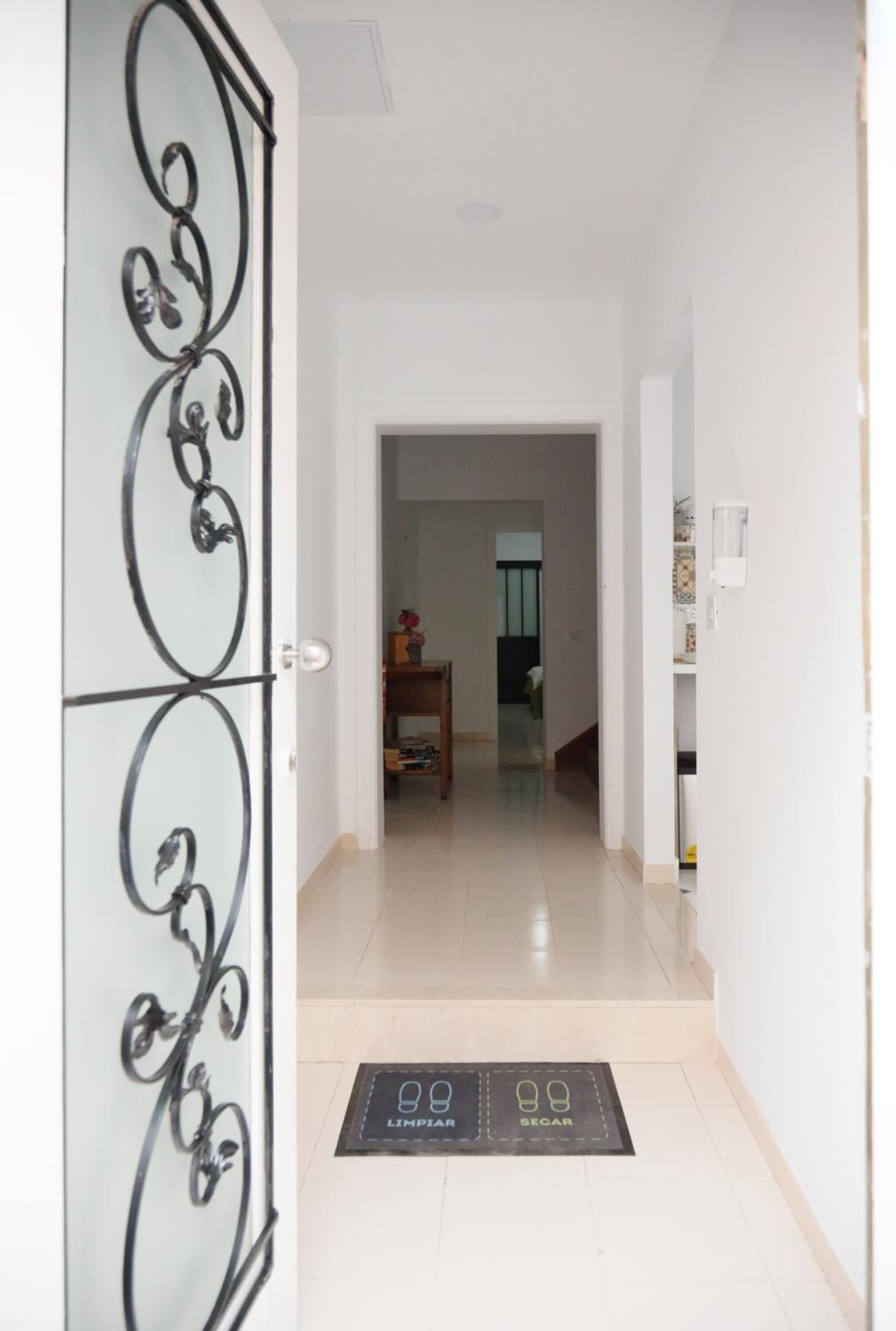 Un pasillo con una puerta blanca que presenta un diseño decorativo de metal. Un felpudo negro con iconos que indican limpieza y seguridad está en el suelo.