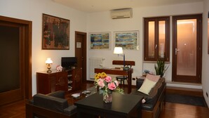 Een woonkamer met een bank, een salontafel, een televisie en een boekenkast. Twee schilderijen hangen aan de muur en er is een raam met uitzicht.