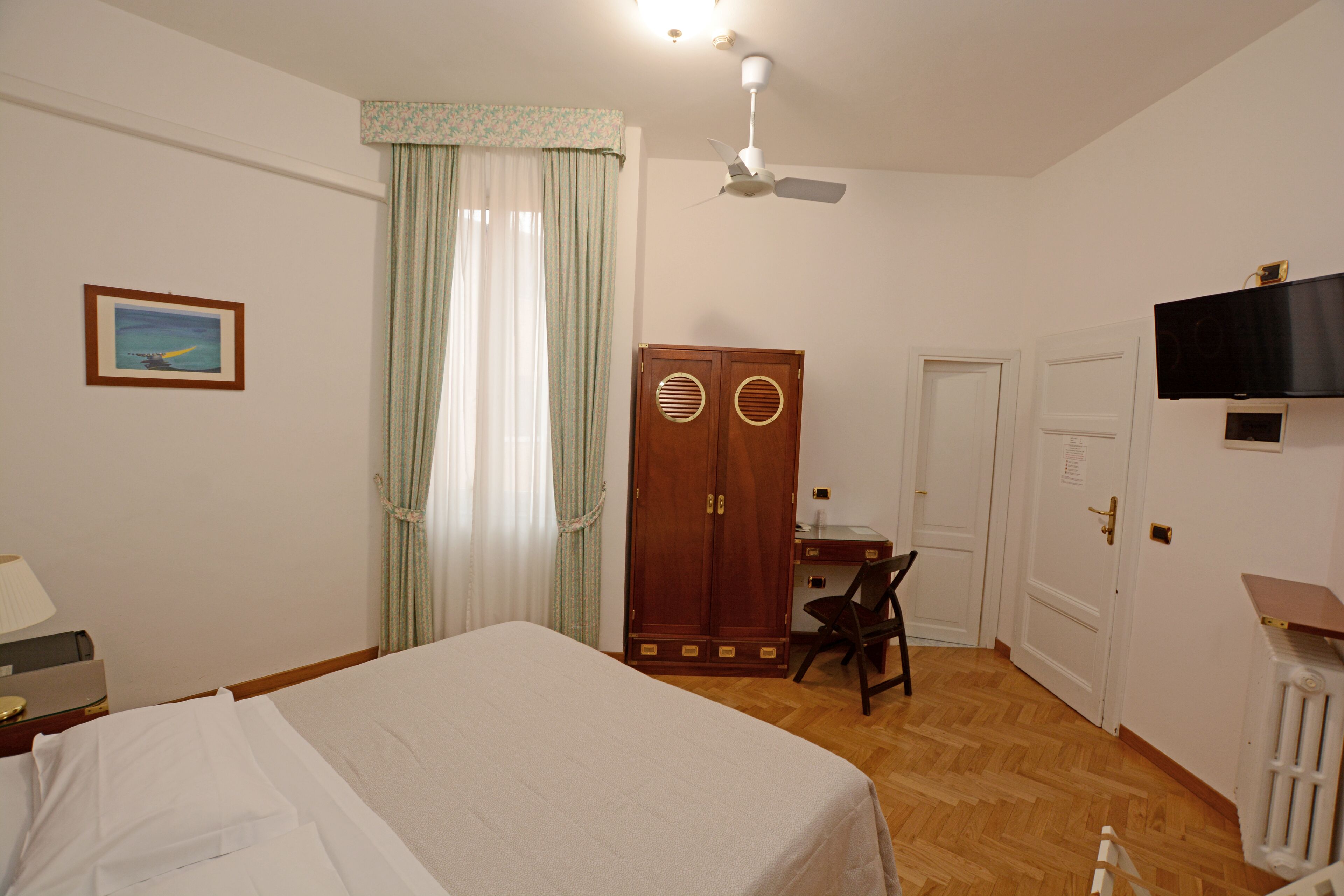 A hotel room with a bed, a desk, a chair, a wardrobe, a television, and a ceiling fan.