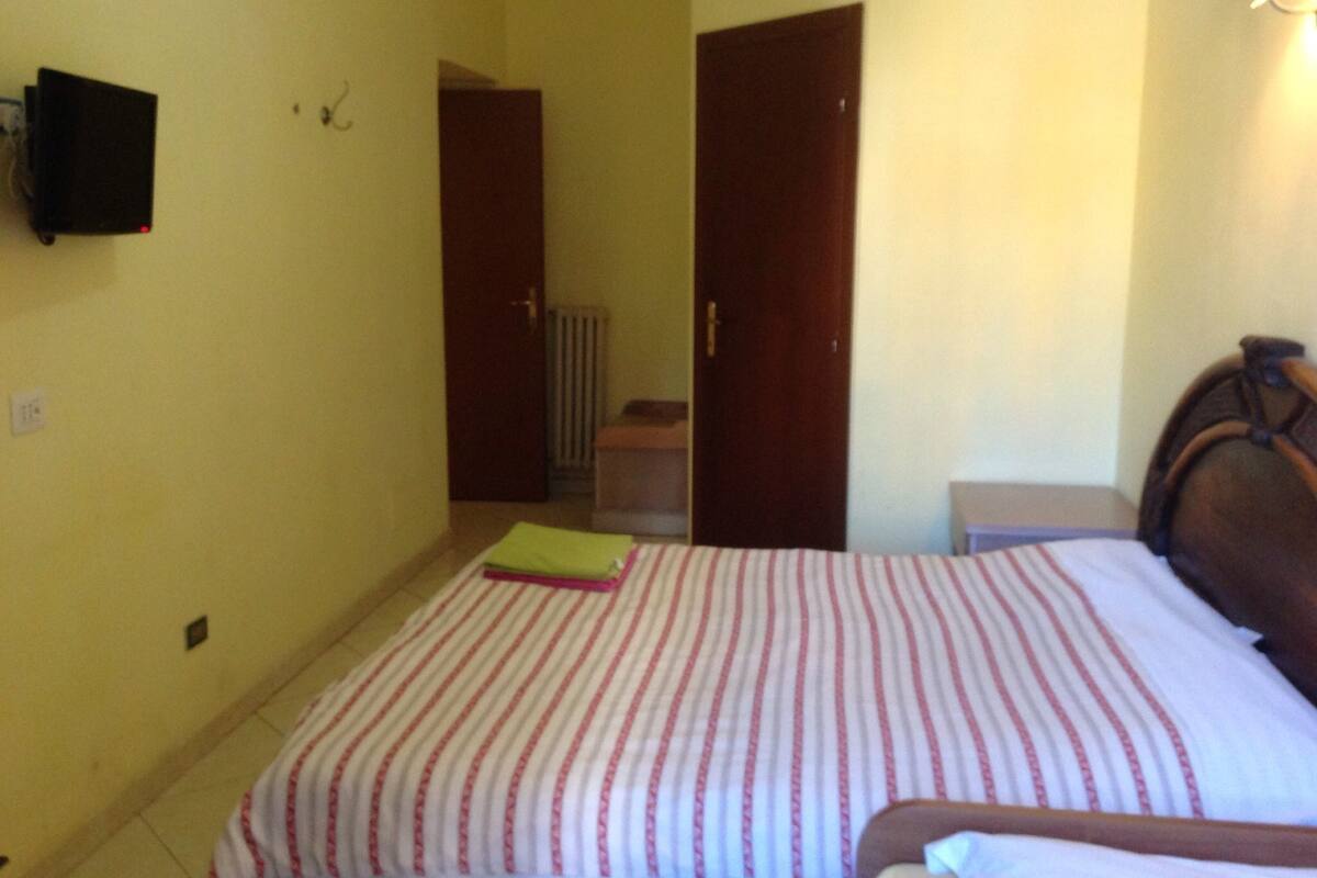 A bedroom with a bed, a bedside table, a wall-mounted TV, and a door leading to another room.