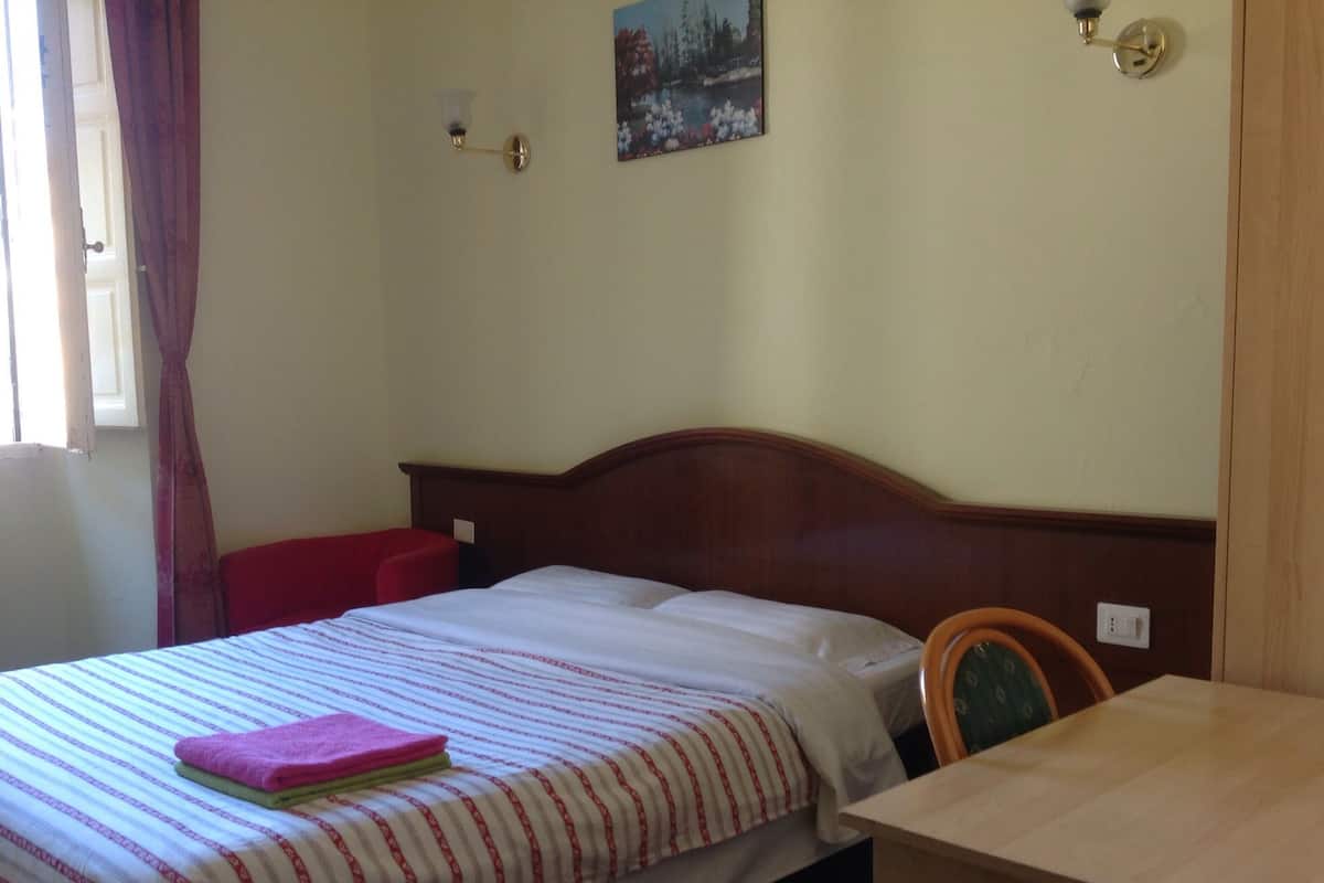 A single bed with a striped bedspread, a red pillow, and a folded pink towel on the bed. There is a wooden nightstand with a lamp, a chair, and a wooden desk with a chair. The room has a window with curtains, a wall-mounted light fixture, and a painting above the bed.