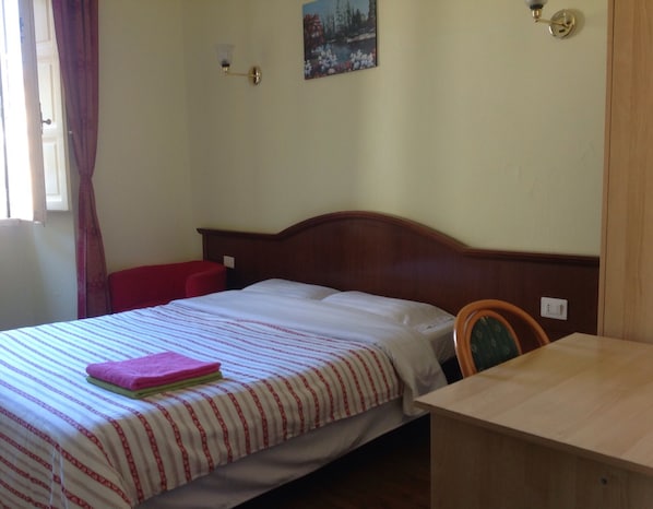 A single bed with a striped bedspread, a red pillow, and a folded pink towel on the bed. There is a wooden nightstand with a lamp, a chair, and a wooden desk with a chair. The room has a window with curtains, a wall-mounted light fixture, and a painting above the bed. - Vittoria & Botel Inn (Rome)