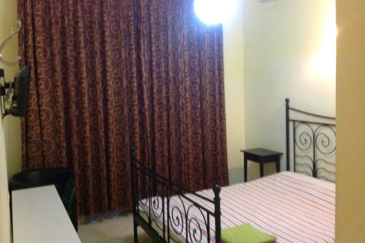 A bedroom with a bed, a desk, a chair, a curtain, and an air conditioning unit.