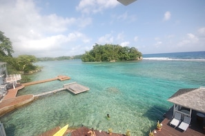 A clear view of a tropical beach resort with a wooden dock extending into the turquoise water, a small island with dense greenery in the background, and a building with a covered seating area on the right. - San Bar 6BR by Jamaican Treasures (Port Antonio)