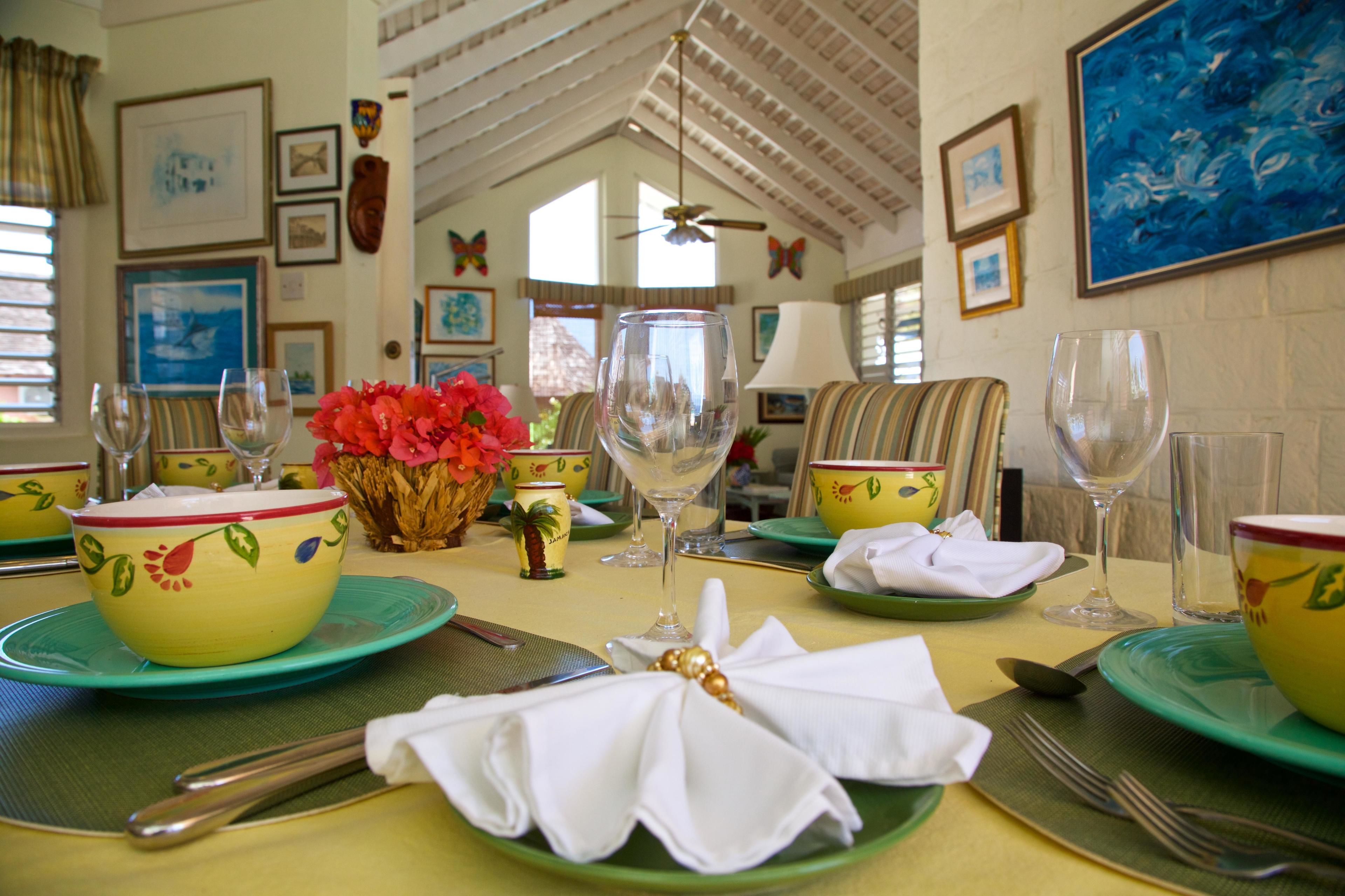 A dining table set with green plates, yellow bowls, and white napkins. There are wine glasses and a vase with red flowers.