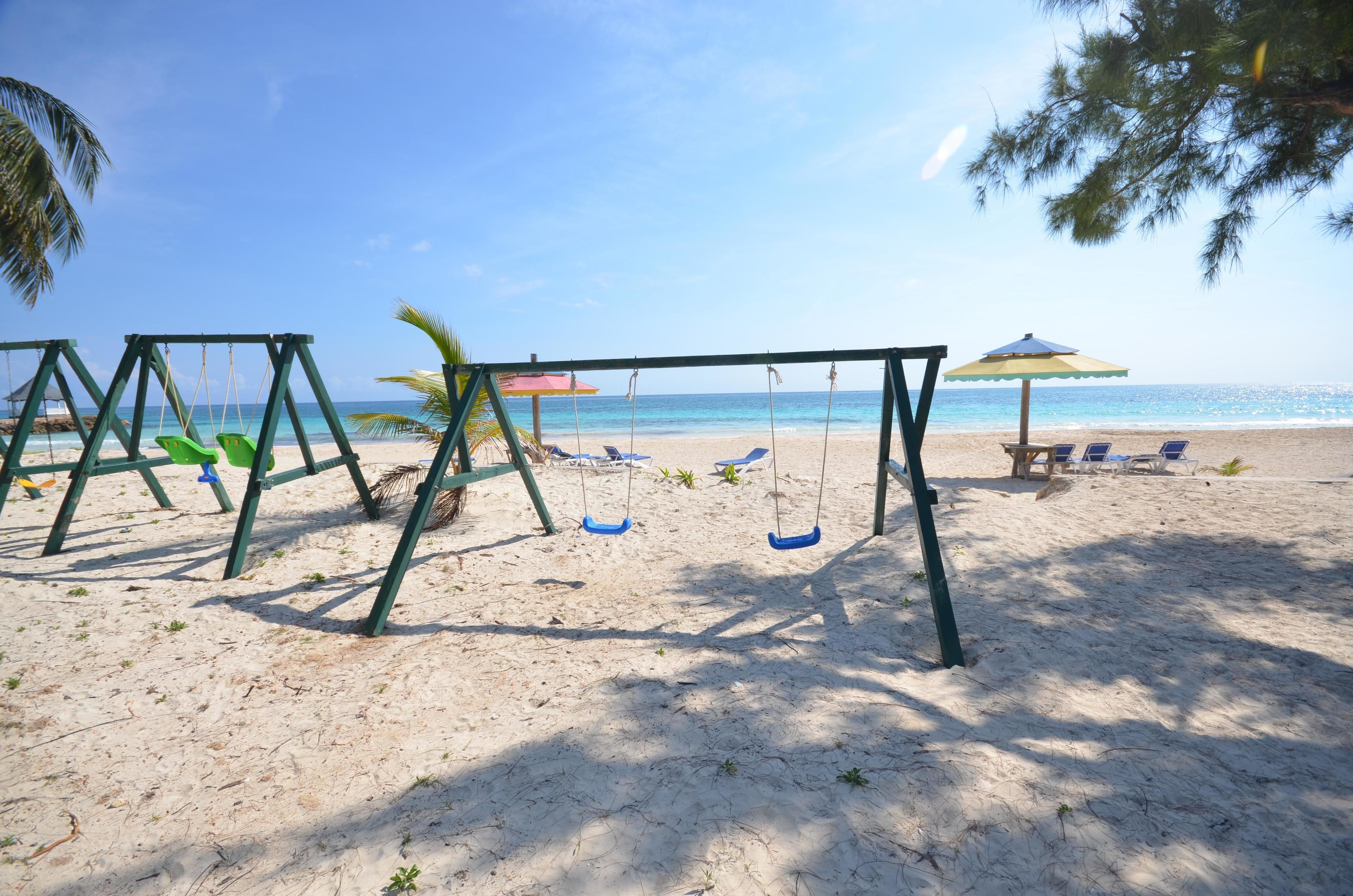 A beach with a swing set, a beach umbrella, and lounge chairs.