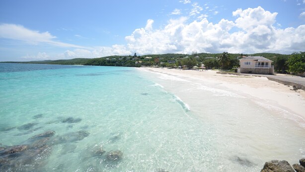 Une plage à l’eau turquoise et limpide, un rivage sablonneux et un petit bâtiment doté d’un balcon.