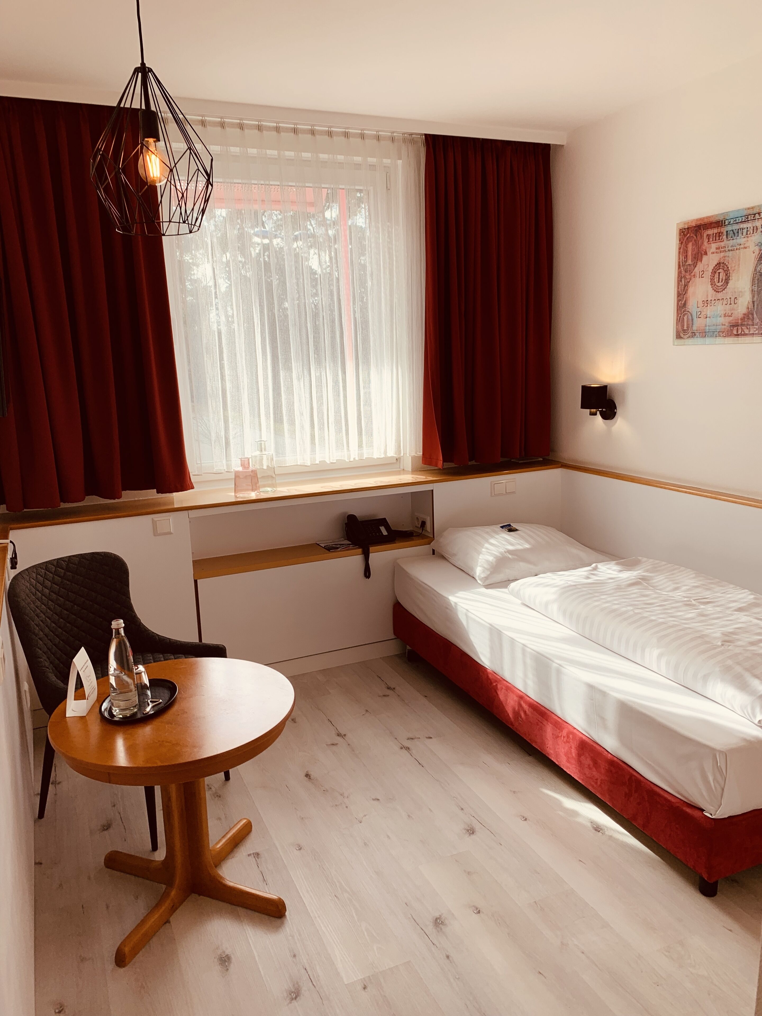 Une chambre d’hôtel de taille réduite, comprenant un lit simple, une petite table avec une chaise, un bureau avec un téléphone et une fenêtre avec des rideaux.