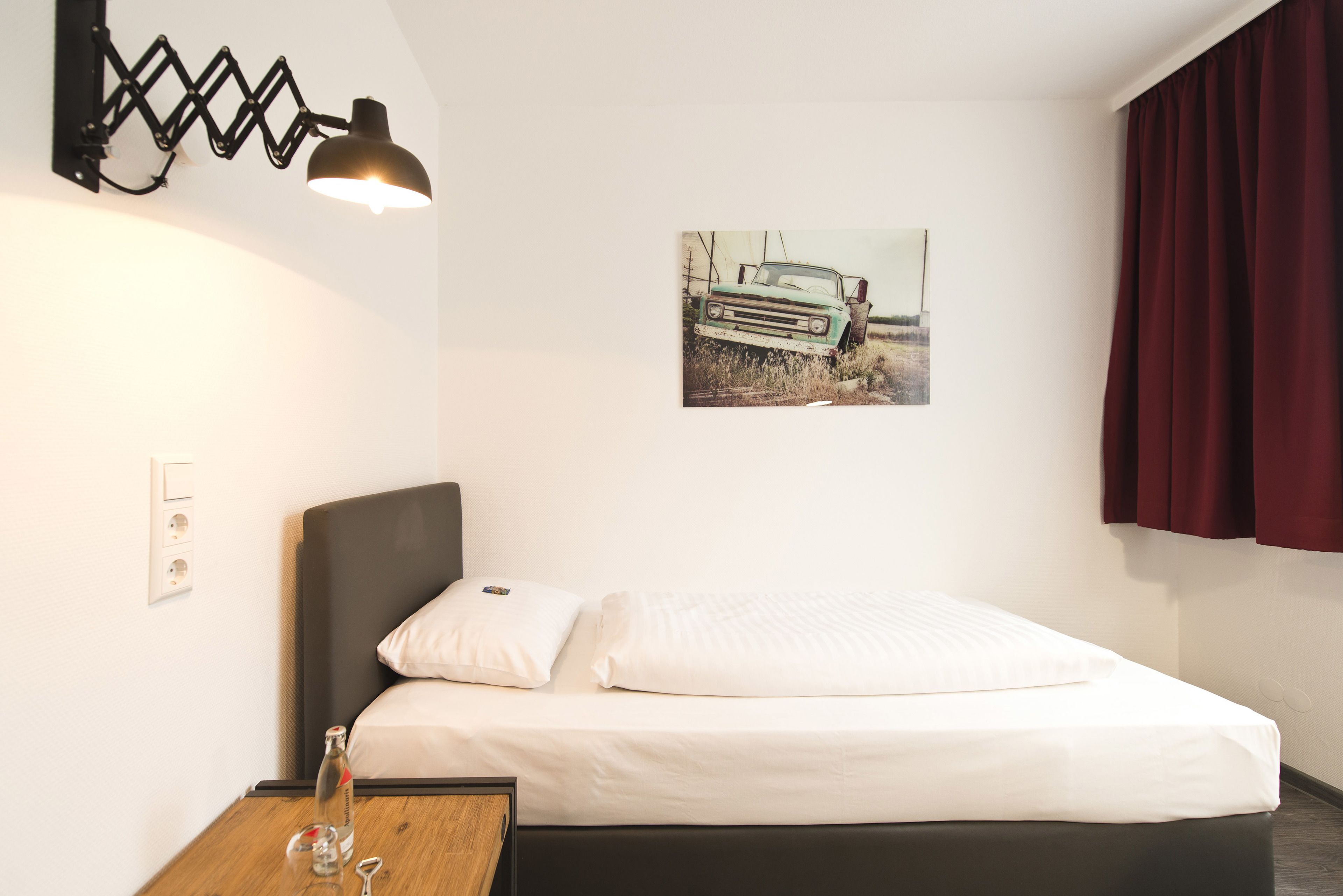 A neatly made bed with white linens, a wall-mounted lamp, a framed picture of a vintage car, and a wooden nightstand with a bottle and glass.