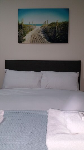 Un dormitorio con cama, almohadas, una colcha y un cuadro de un camino en la playa en la pared.
