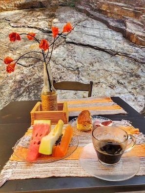 A table set with a vase of red flowers, a plate of fruit, a pastry, and a cup of coffee on a textured placemat. - Pousada Marotta (Ouro Preto)