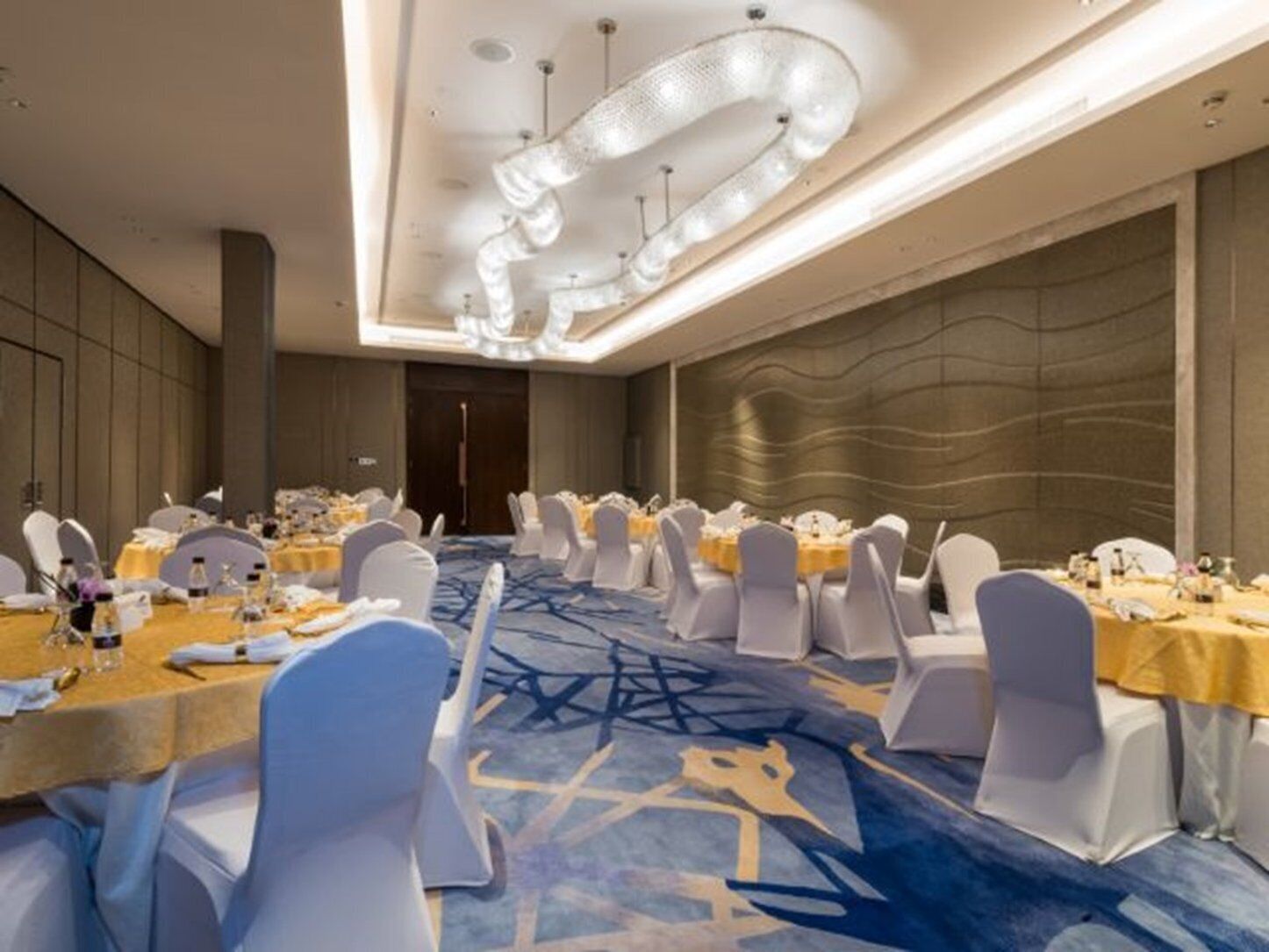 A banquet hall with round tables, white chairs, and a blue carpet with geometric patterns.