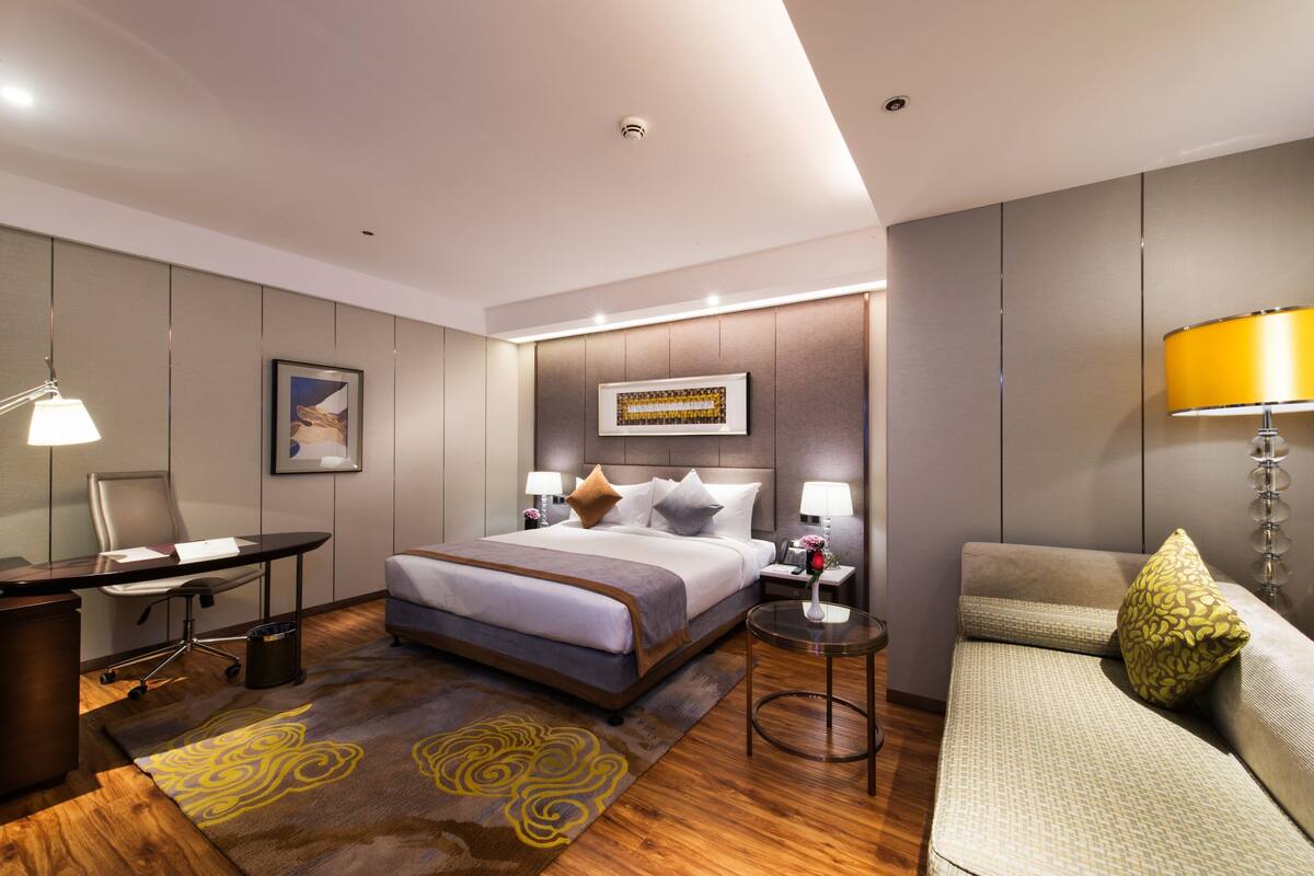 A modern hotel room with a large bed, a desk, a chair, a sofa, and a small table.