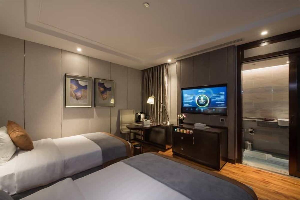 A modern hotel room with two beds, a flat-screen TV, a desk with a lamp, and a minibar.