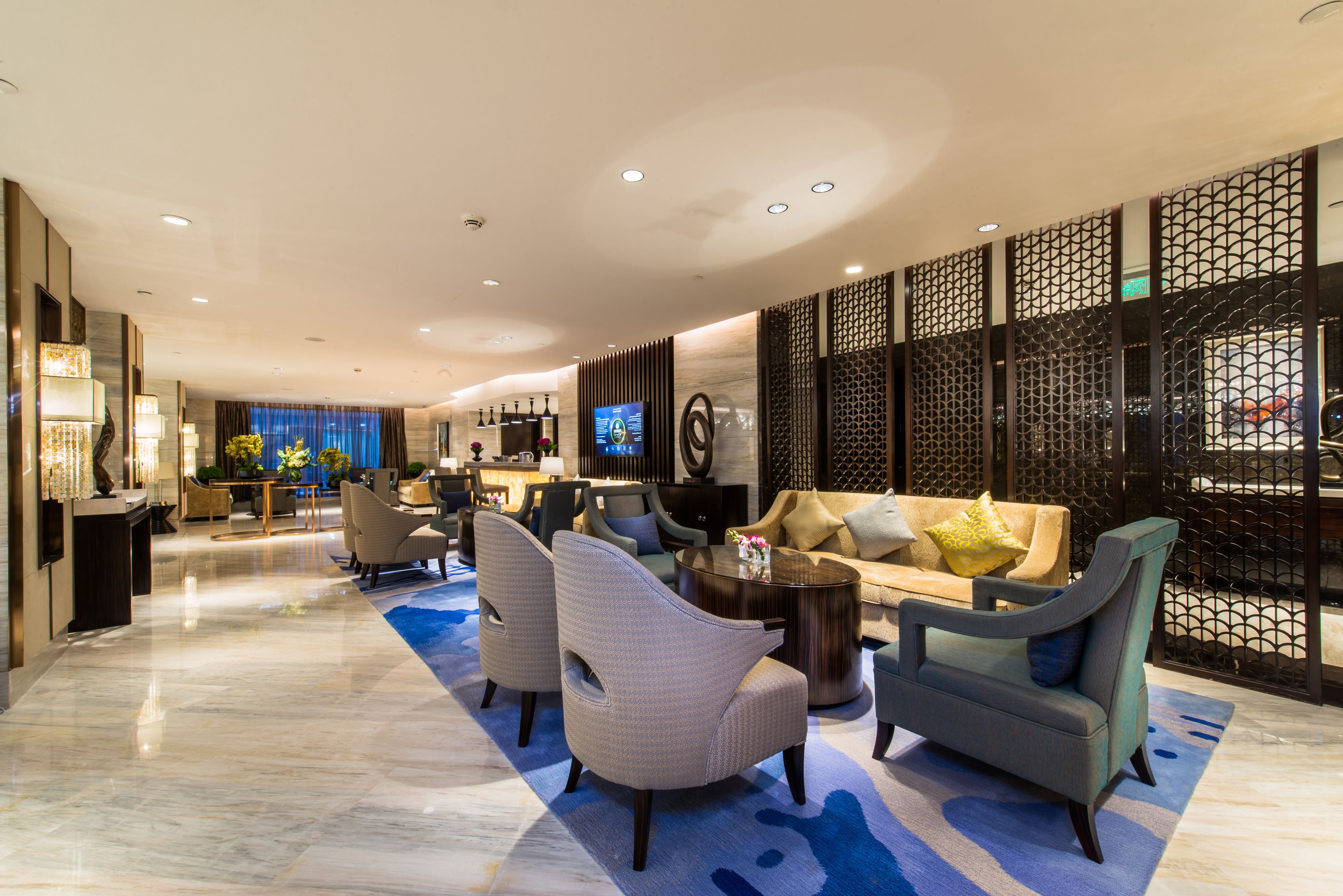 A modern hotel lobby with a reception desk, seating area with sofas and chairs, a TV screen, and decorative elements.