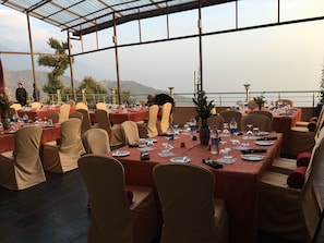 An outdoor dining area with tables set for guests, surrounded by a glass enclosure with a view of the mountains. - Norbu House (Dharamshala)