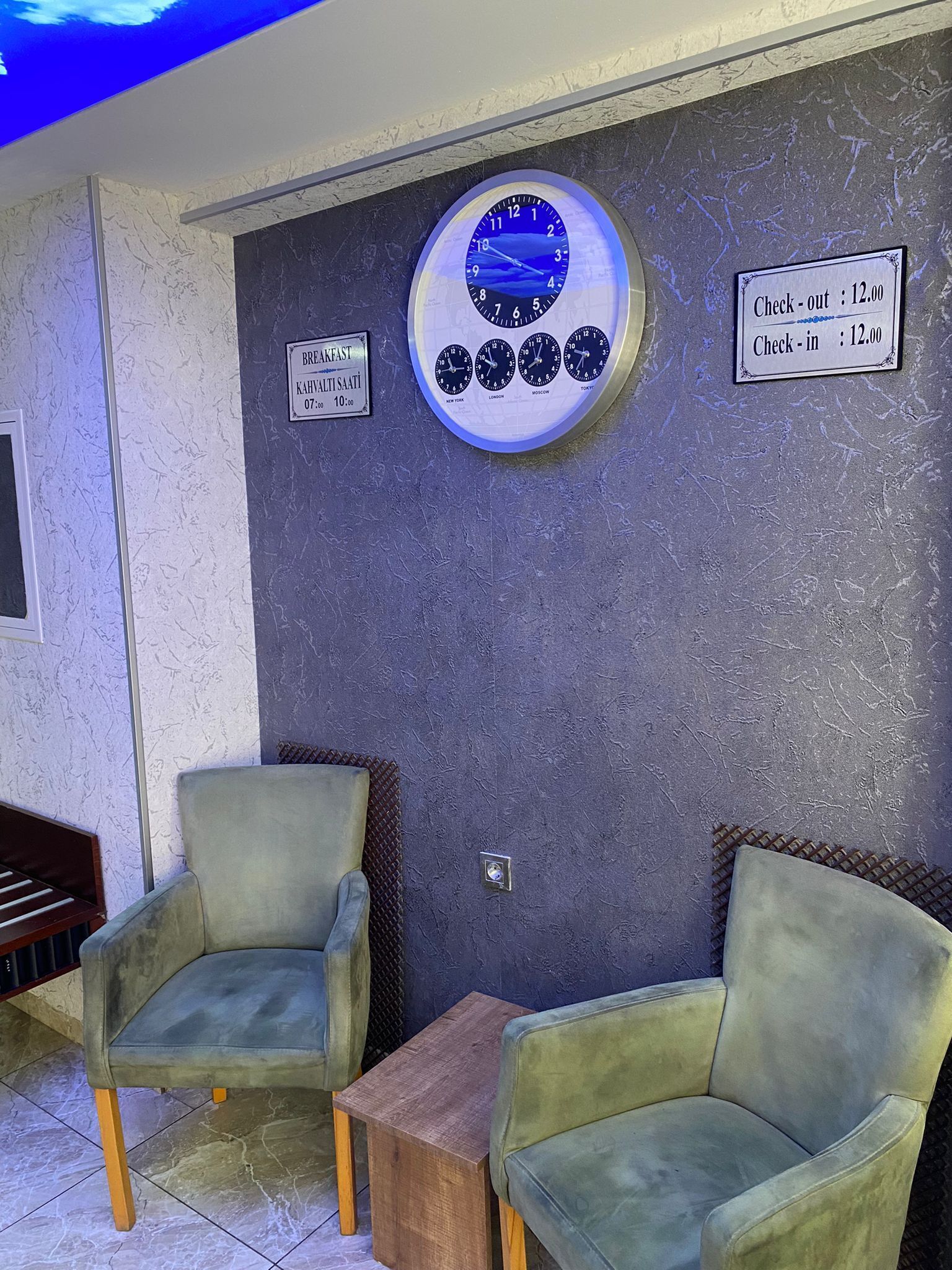 A room with two green armchairs, a wooden table, a wall clock, and signs indicating breakfast hours and check-out/check-in times.