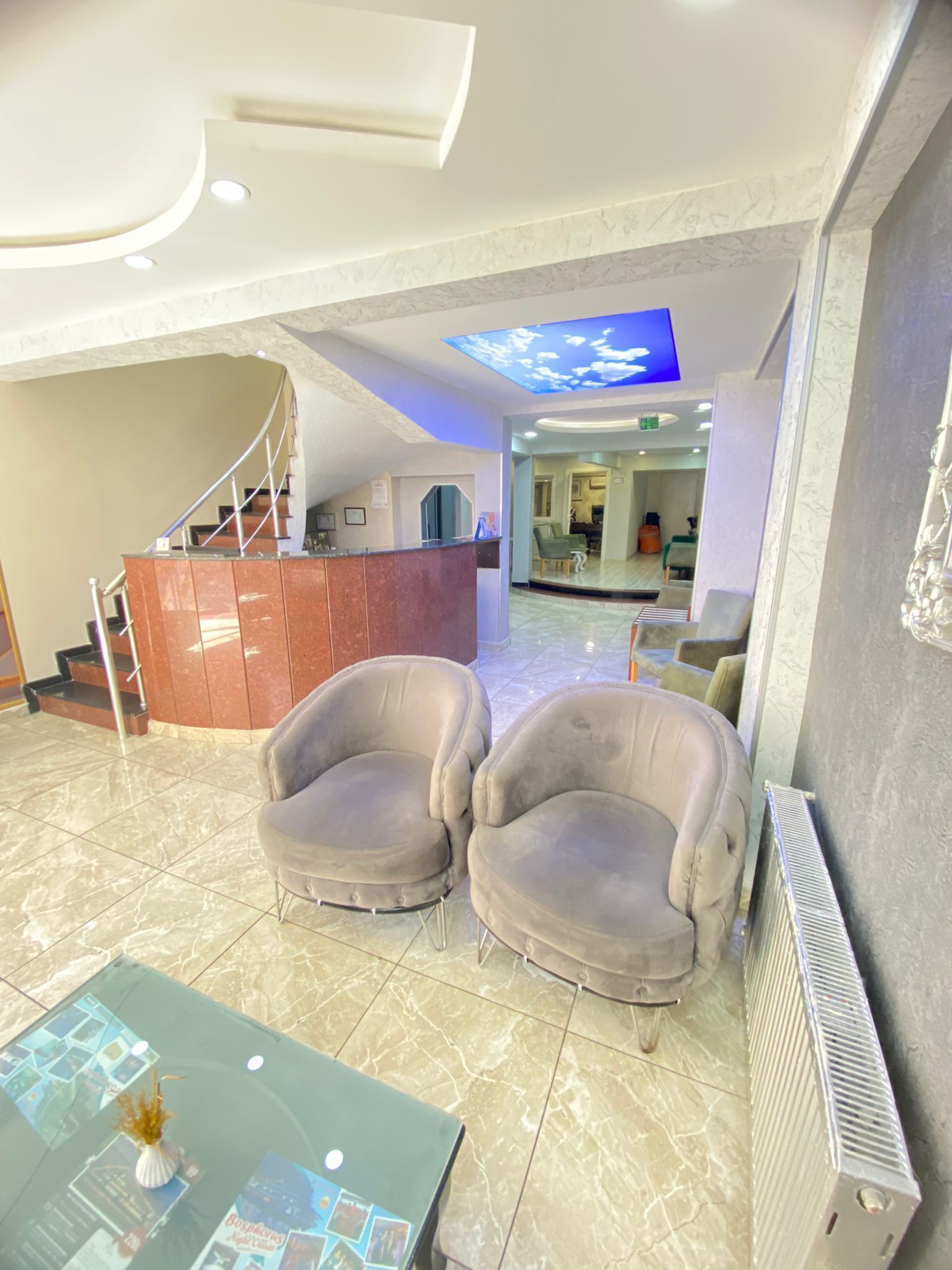 A lobby with a reception desk, two armchairs, a glass coffee table, and a staircase.