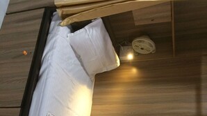 A compact, well-lit room with a wooden floor, a bed with a white mattress and a brown blanket, and a small fan on the ceiling. - Golden Dormitory - Hostel (Mumbai)