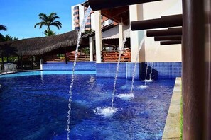 Une piscine avec des jets d’eau, une structure dotée d’un toit de chaume et, au fond, un bâtiment agrémenté de balcons.