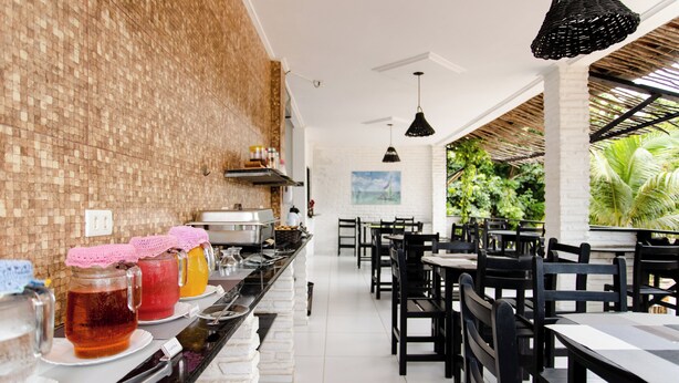 Una larga mesa de buffet con diferentes alimentos y bebidas, rodeada de sillas y mesas negras en un comedor amplio y luminoso con una pared de ladrillo y vistas a la vegetación exterior.