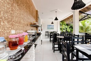 A long buffet table with various food items and drinks, surrounded by black chairs and tables in a bright, open dining area with a brick wall and a view of greenery outside.