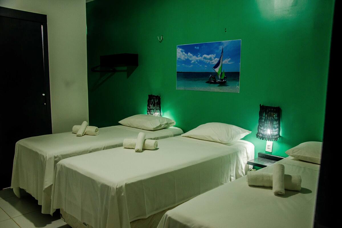 A hotel room with two single beds, a green wall, a painting of a sailboat, and a small table with a lamp.