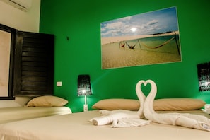 Una habitación de hotel con pared verde, una cama con ropa de cama color crema y un cuadro enmarcado con una playa, además de dos toallas dispuestas en forma de corazón.