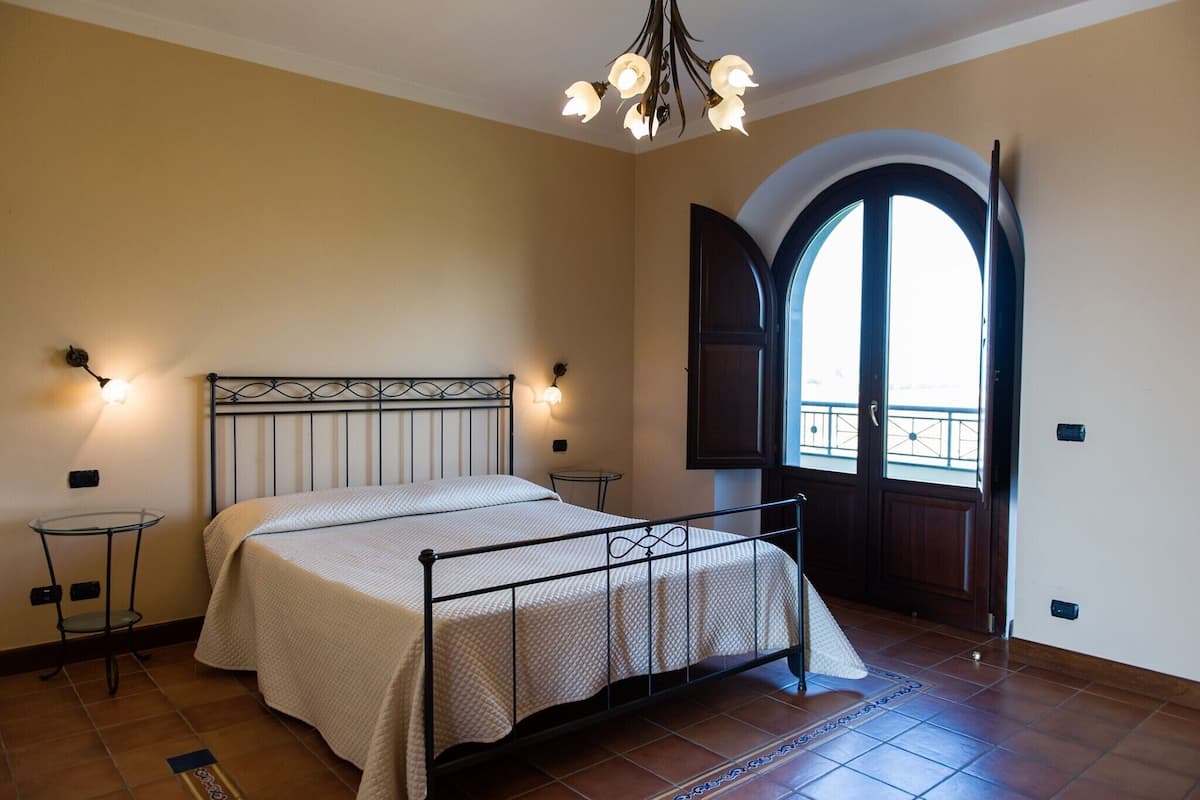 Ein Schlafzimmer mit einem Bett, zwei Nachttischen, einer Kristallleuchter und einem großen, bogenförmigen Fenster.