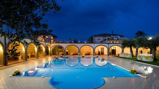 A large outdoor swimming pool with a tiled deck, surrounded by arched structures and a building in the background. - La Vecchia Torre (San Nicola Manfredi)