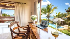 A wooden desk with a laptop, a vase with flowers, and a view of a pool and palm trees. - Residence Pé na Areia (Porto Seguro)
