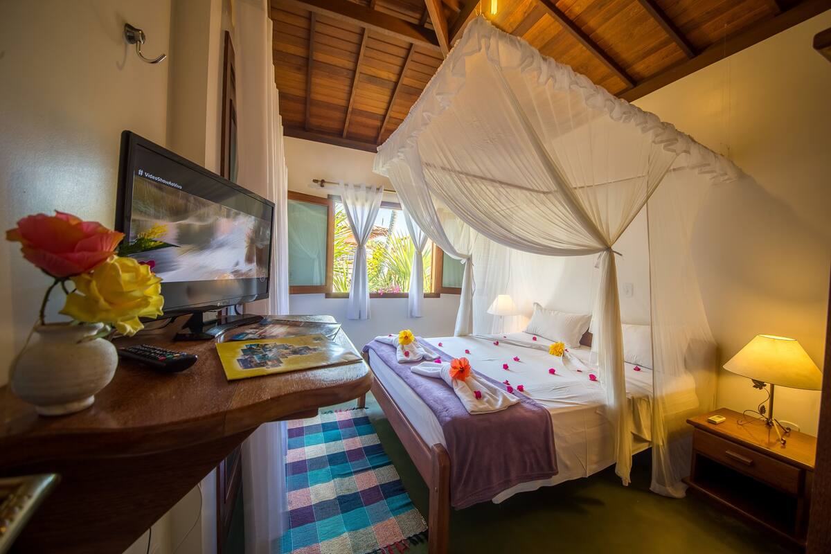 Un dormitorio con una cama, un televisor, un escritorio con un florero y una mesita de noche con una lámpara.