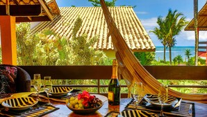 A dining setup with a table set for two, a bottle of wine, and a bowl of fruit. The table is outdoors with a view of a tiled roof and palm trees. - Residence Pé na Areia (Porto Seguro)