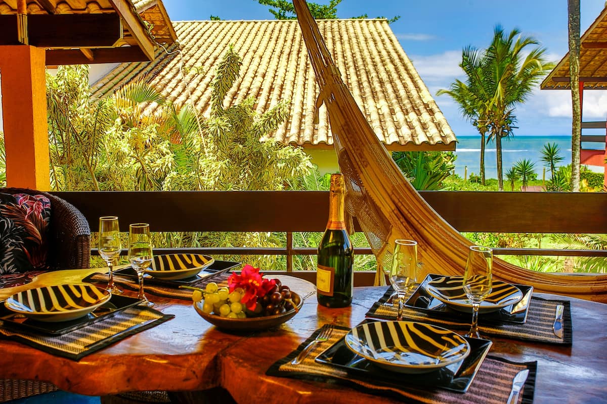 Una mesa para dos, con una botella de vino y un cuenco de fruta. La mesa está al aire libre, con vistas a un tejado de azulejos y palmeras.