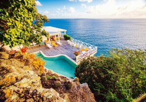 A cliffside pool with a wooden deck, lounge chairs, and a view of the ocean.