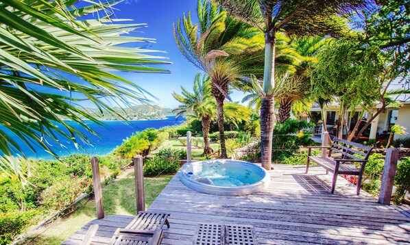 A wooden deck with a hot tub, surrounded by lush greenery and a view of the ocean.