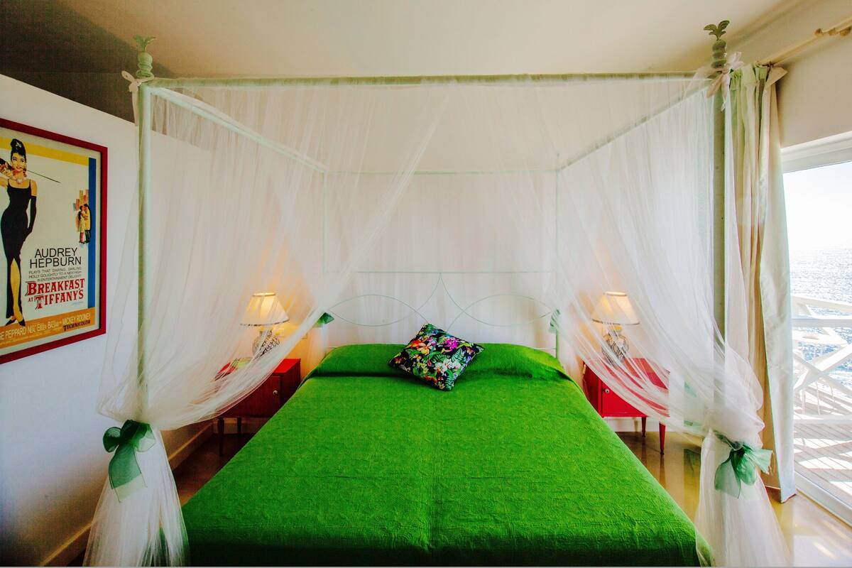 A room with a four-poster bed, green bedding, and a floral pillow. There are two bedside lamps and a poster on the wall.