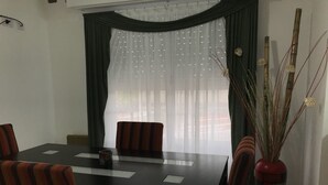 Uma área de refeições com mesa e cadeiras, um vaso com plantas secas e uma janela com cortinas leves.