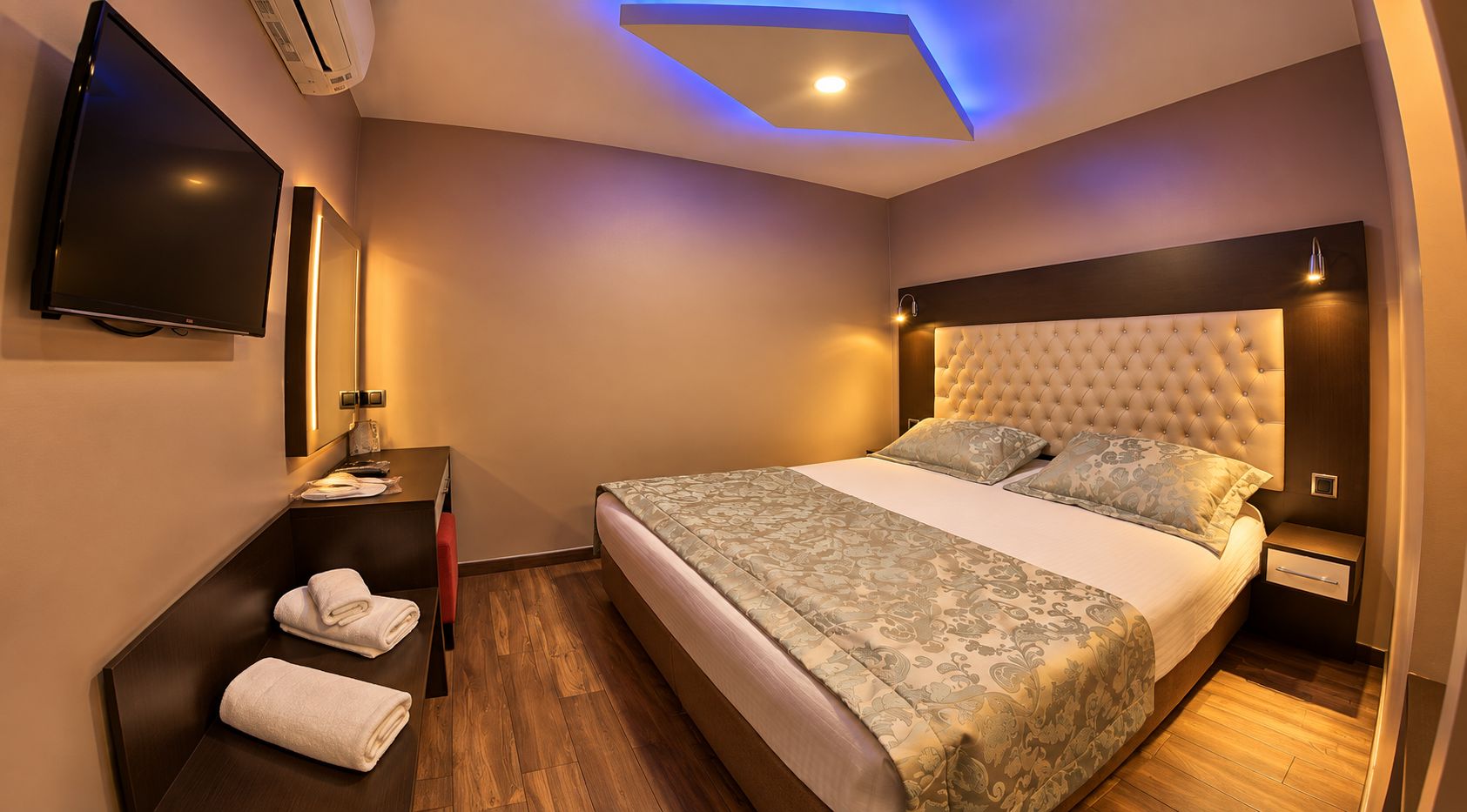 A modern hotel room with a large bed, a flat-screen TV, a bedside table with a lamp, and a wall-mounted air conditioner.
