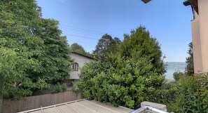 Une vue d’une maison avec une terrasse en bois, une végétation luxuriante et un plan d’eau en arrière-plan.